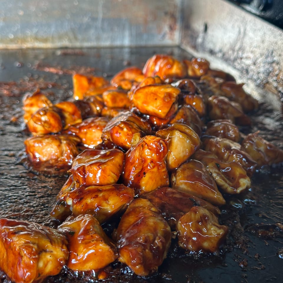 Glossy, sauced chicken pieces cooking on a dark, well-used griddle.