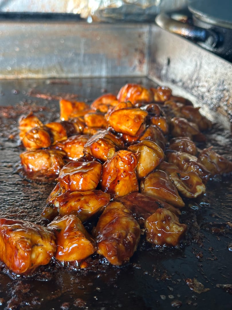 Glossy, sauced chicken pieces cooking on a dark, well-used griddle.