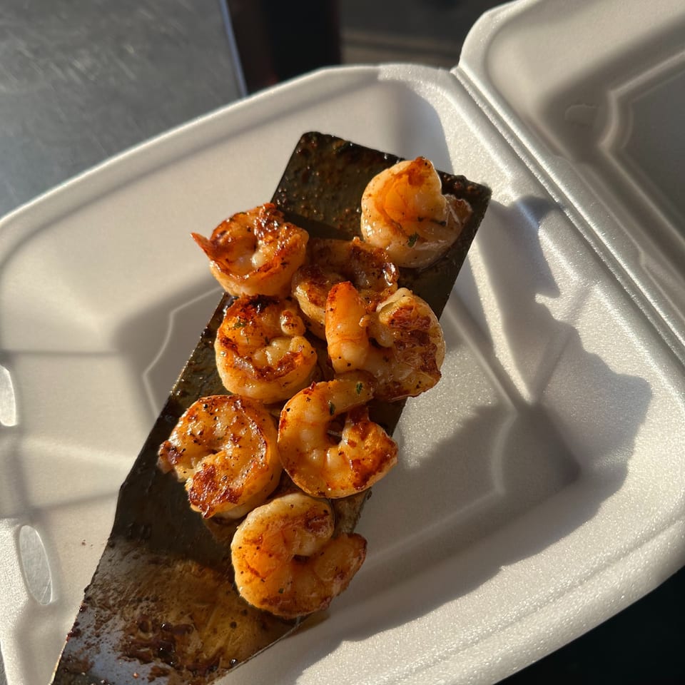 Grilled seasoned shrimp on a spatula over a white takeout container.