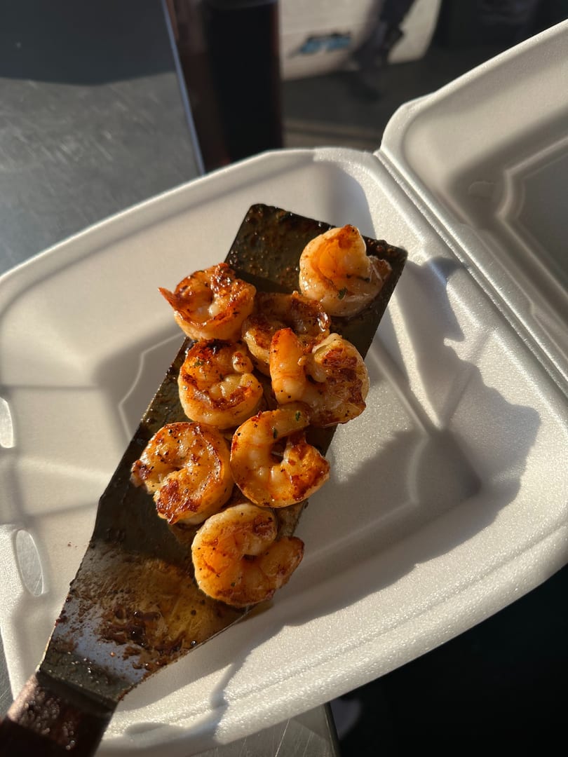 Grilled seasoned shrimp on a spatula over a white takeout container.