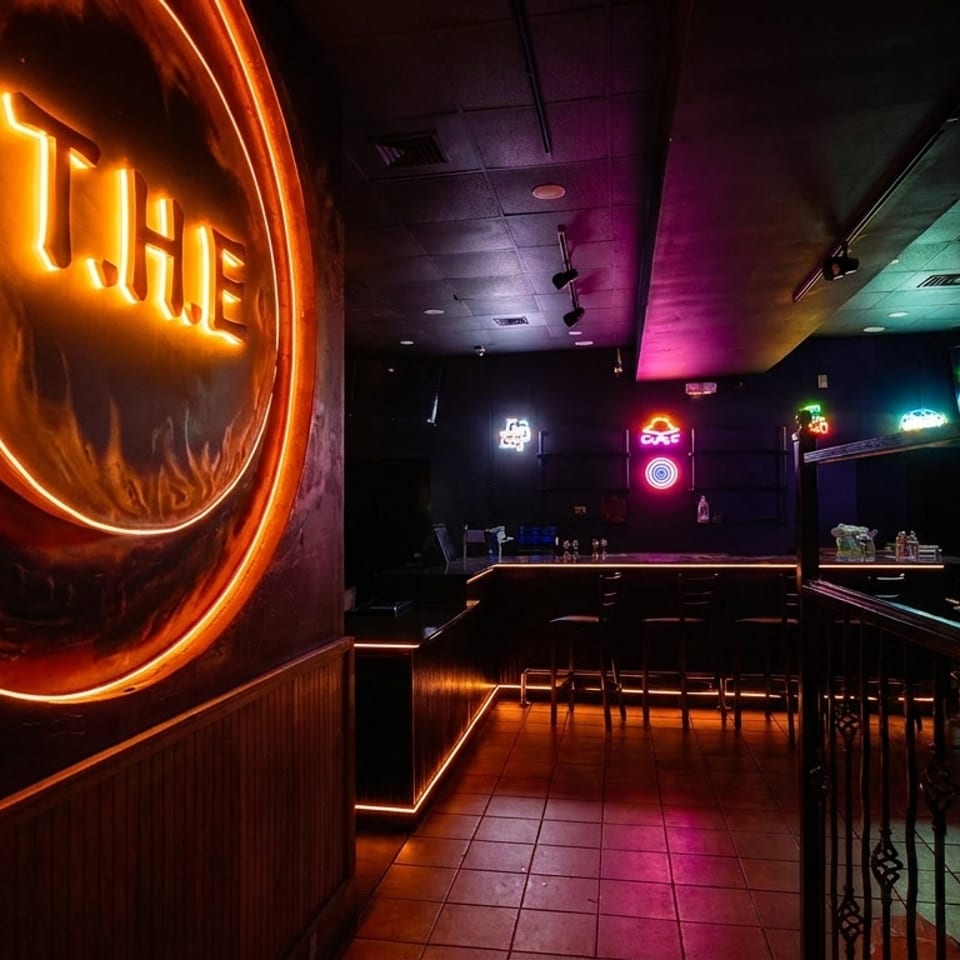 Dark bar interior with orange neon "THE" sign, colorful neon lights, and empty bar stools.
