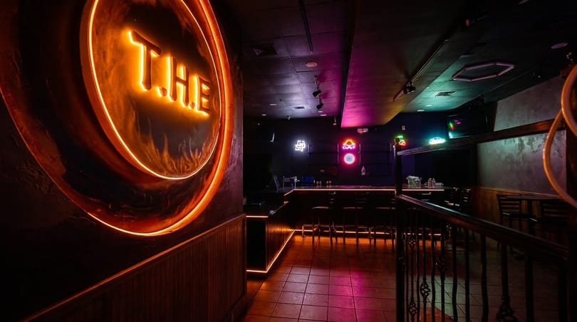 Dark bar interior with orange neon "THE" sign, colorful neon lights, and a tiled floor.