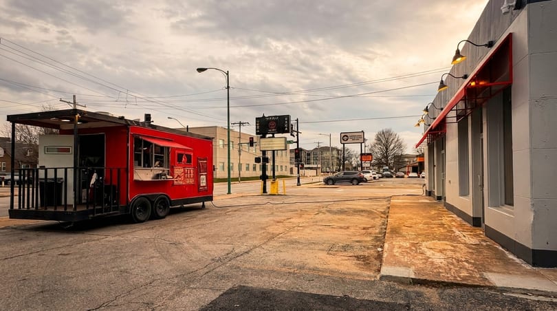 A red food truck with an open serving window parked on a street next to a building with red awnings.