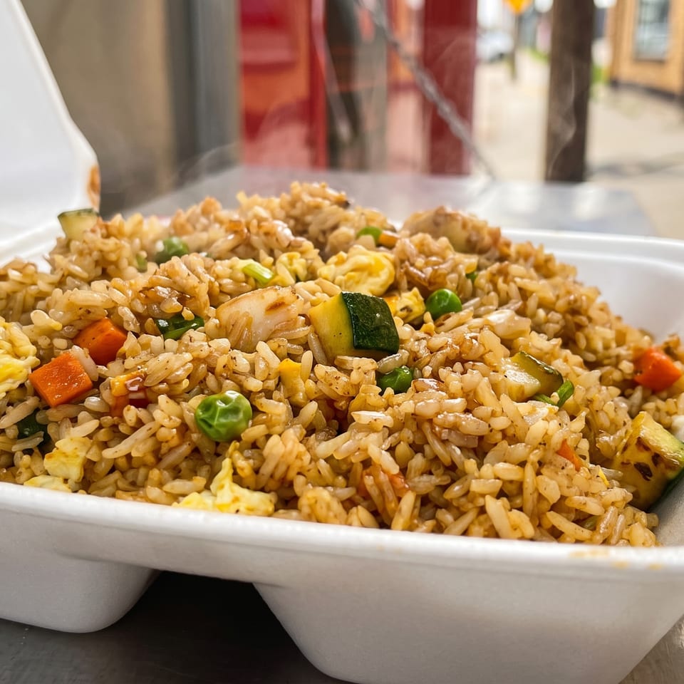 Steaming hot fried rice with chicken, zucchini, carrots, peas, and egg in a white takeout container.