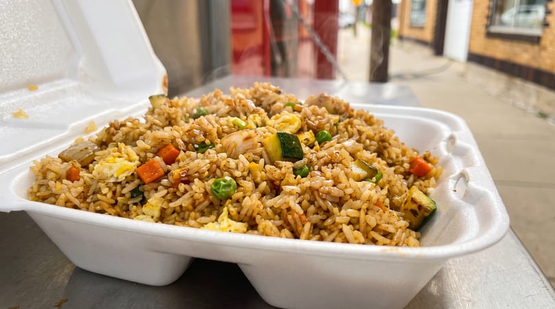 Steaming hot fried rice with chicken, zucchini, carrots, peas, and egg in a white takeout container.