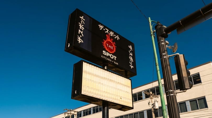 A black sign for "The Spot Kitchen + Bar" with a fiery logo against a clear blue sky.